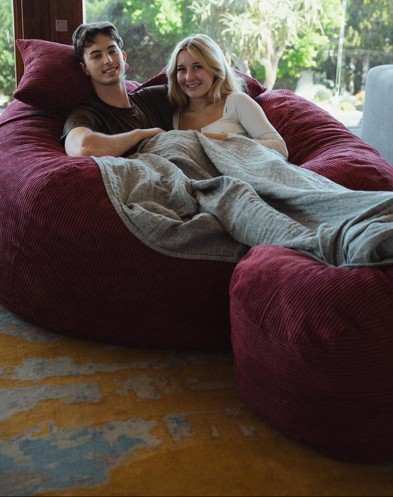 Two people sitting on a large red bean bag chair indoors with a view of greenery outside.