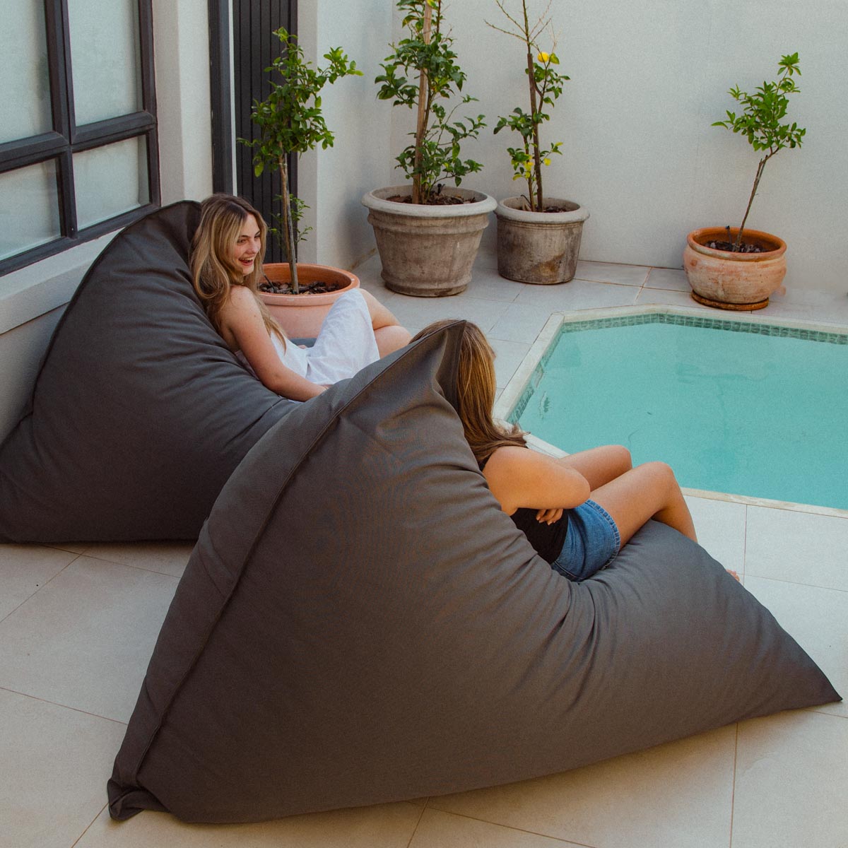 Two people sitting on bean bags by a pool with potted plants around.