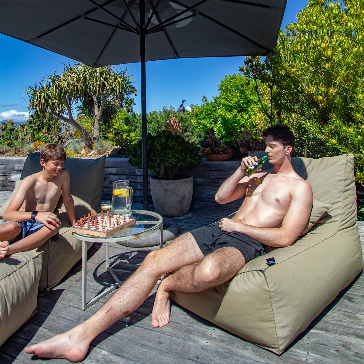 Two men relaxing on outdoor furniture under an umbrella with a chessboard and drinks.