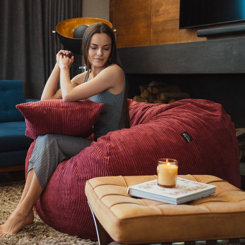Woman sitting on a red bean bag chair in a living room with a blue couch and a candle on an ottoman.