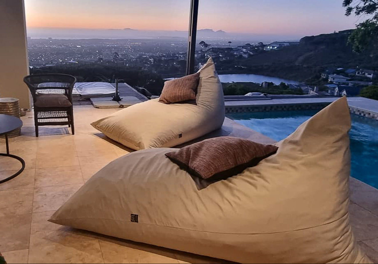 Large beige bean bags with brown cushions by a poolside with a cityscape view at dusk.