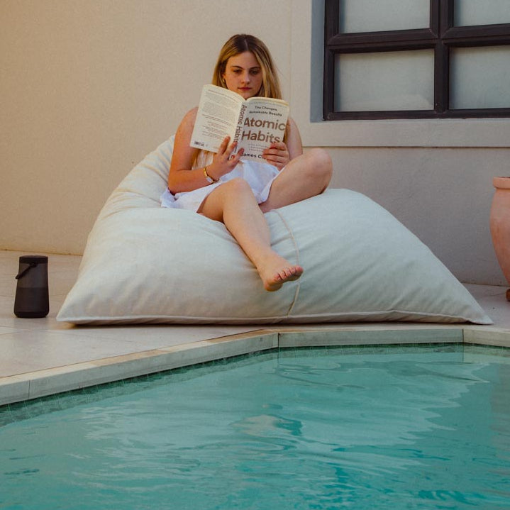 Woman reading a book on a large bean bag by a pool.