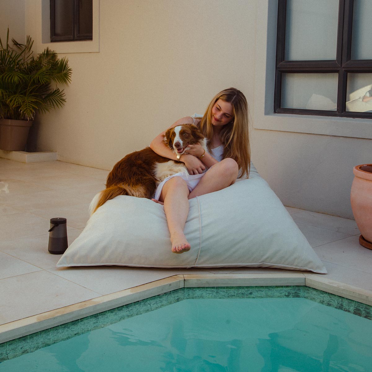Woman sitting on a large pillow by a pool with a dog on her lap