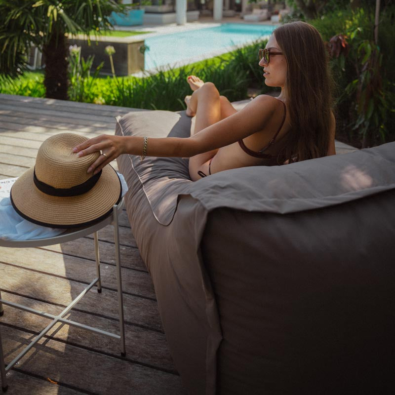Woman relaxing on a lounge chair by a pool with a hat and sunglasses on a table.