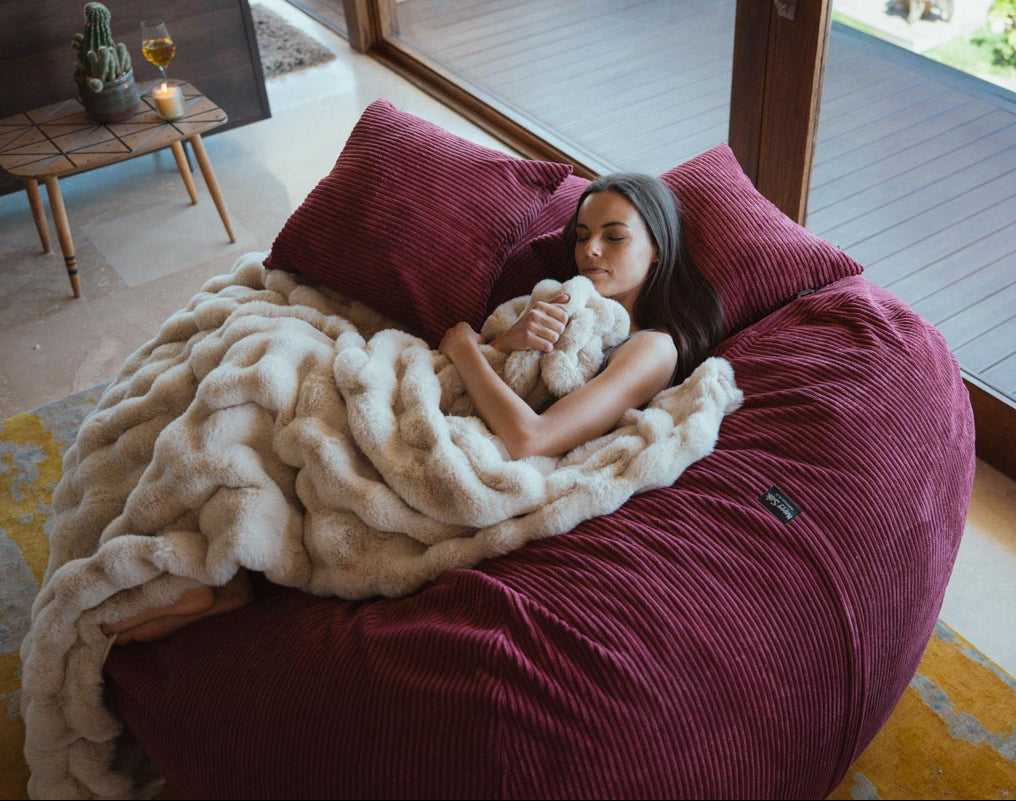 Woman lying on a large burgundy bean bag with a white blanket in a cozy room.