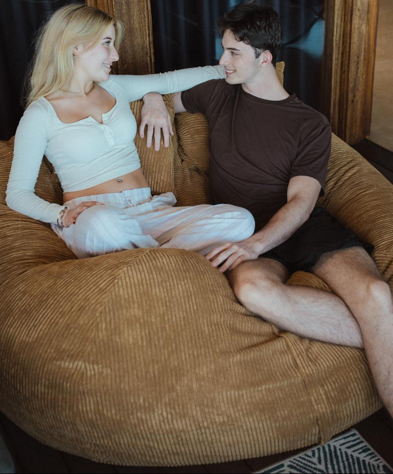 Two people sitting on a brown bean bag chair indoors.