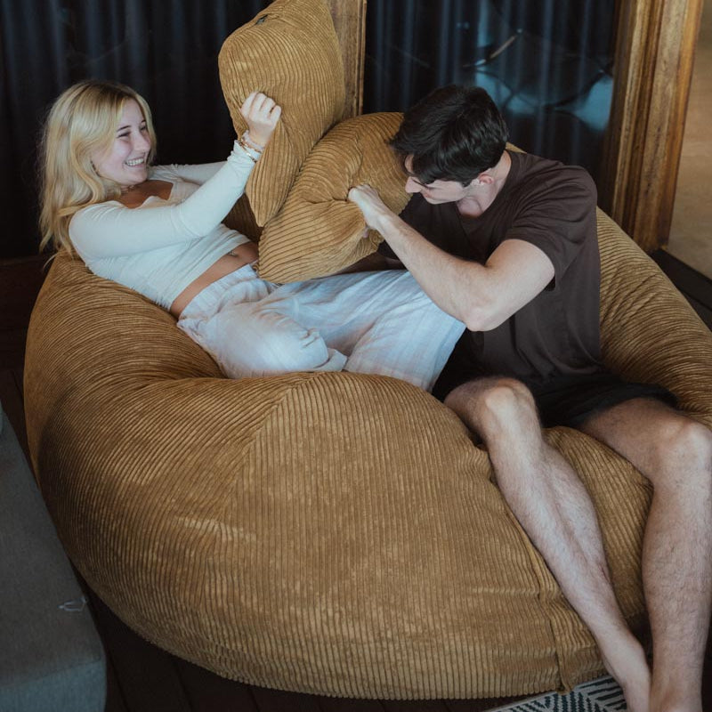 Two people sitting on a large brown bean bag chair indoors.