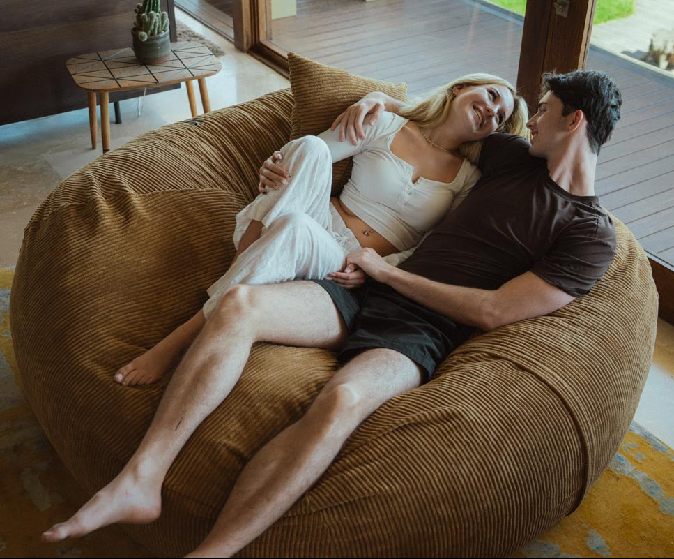 Two people lying on a large brown bean bag chair in a modern living room.