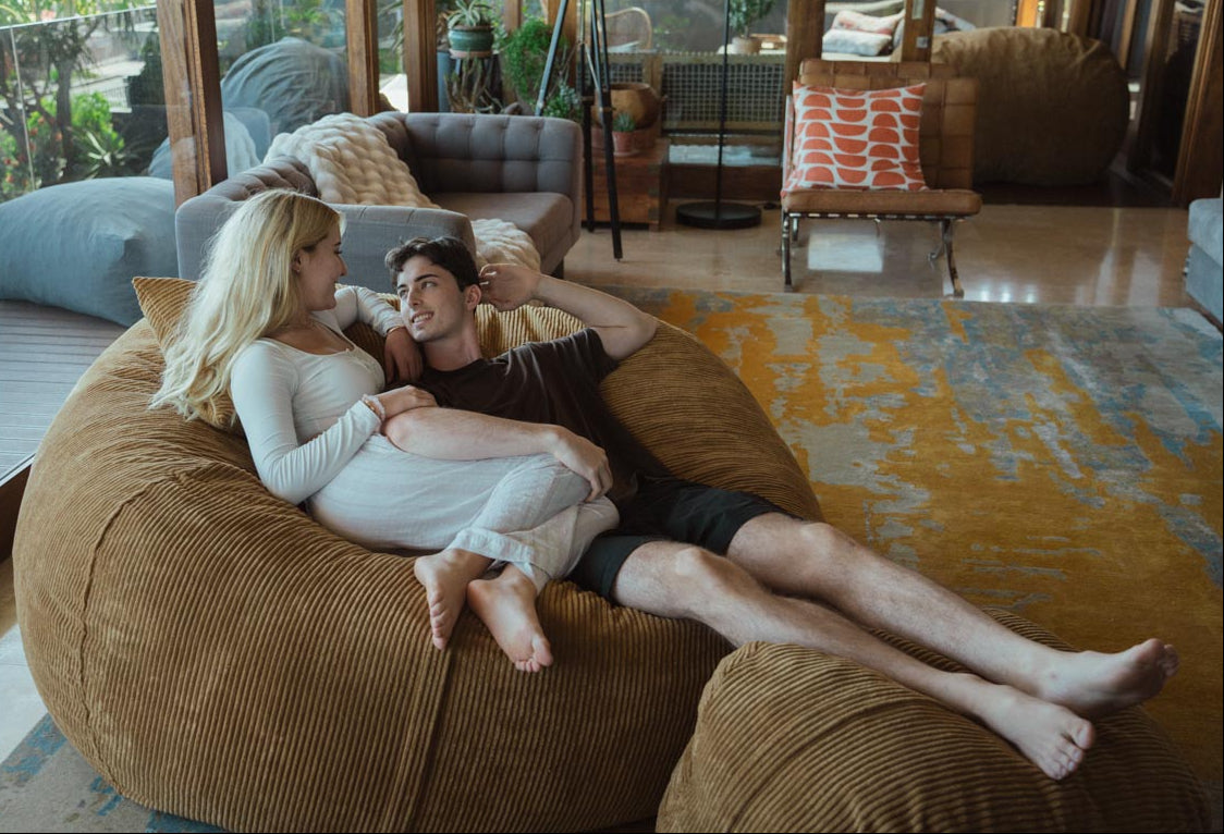Two people sitting on a large brown bean bag in a modern living room.