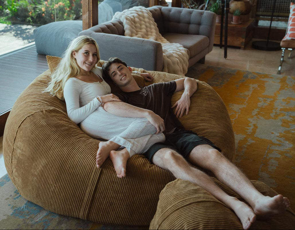 Two people sitting on a large brown bean bag in a living room.
