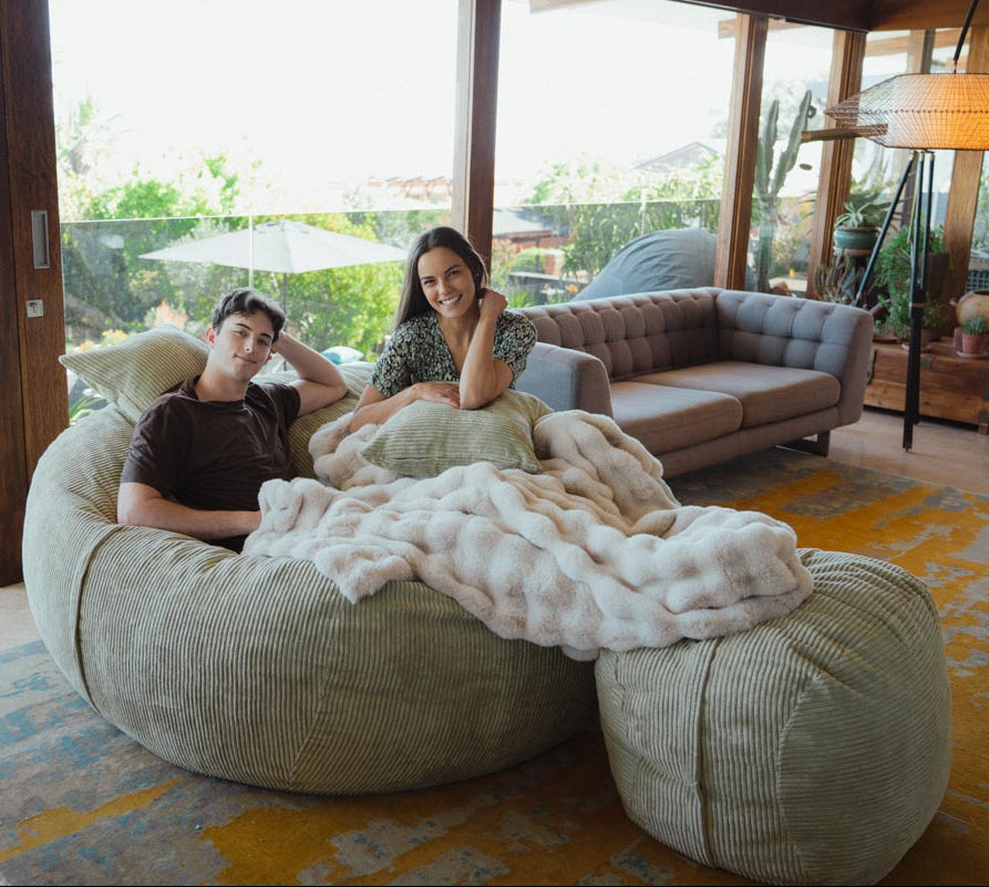 Two people sitting on a large bean bag in a living room with a couch and outdoor view.