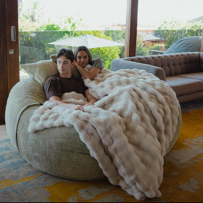 Two people sitting on a large bean bag with a blanket, surrounded by a cozy living room.