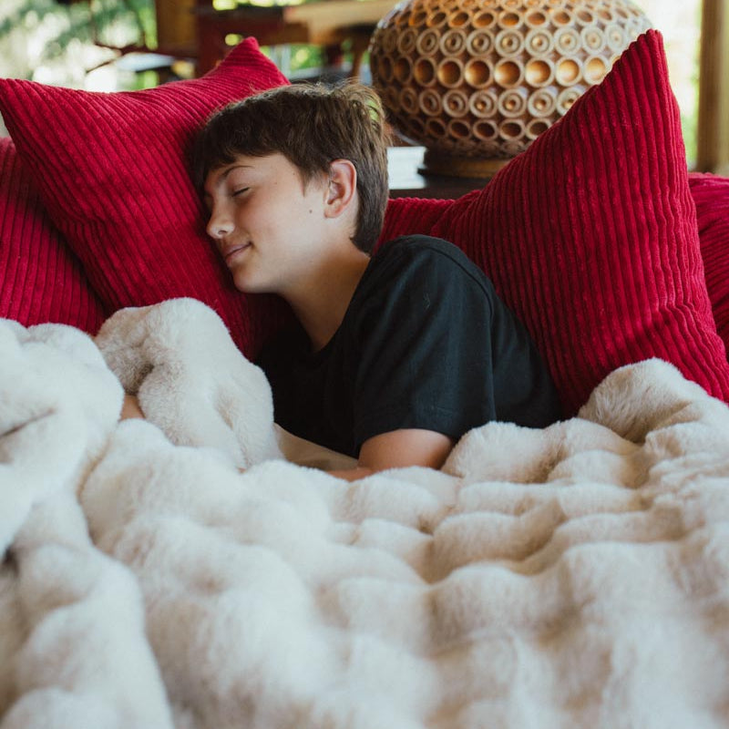 Person lying on a couch with red pillows and a textured blanket, with a lamp in the background.