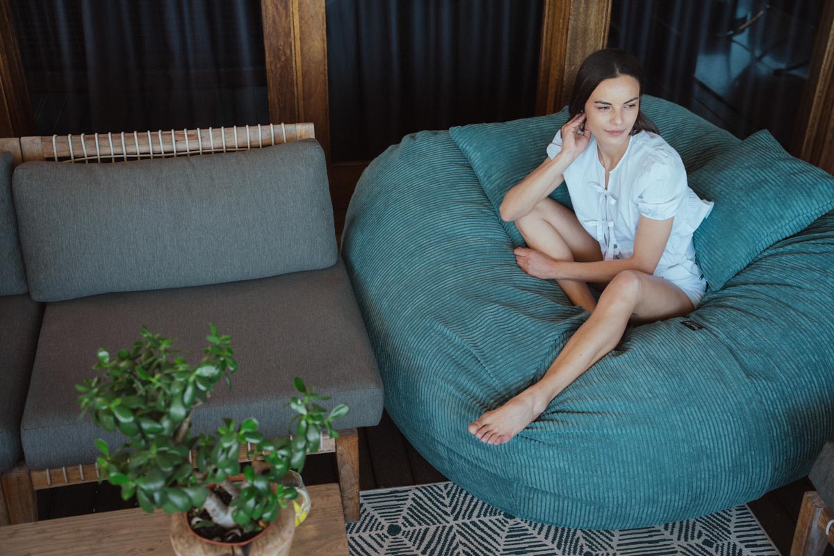 Woman sitting on a teal bean bag chair in a room with a gray sofa and a plant.