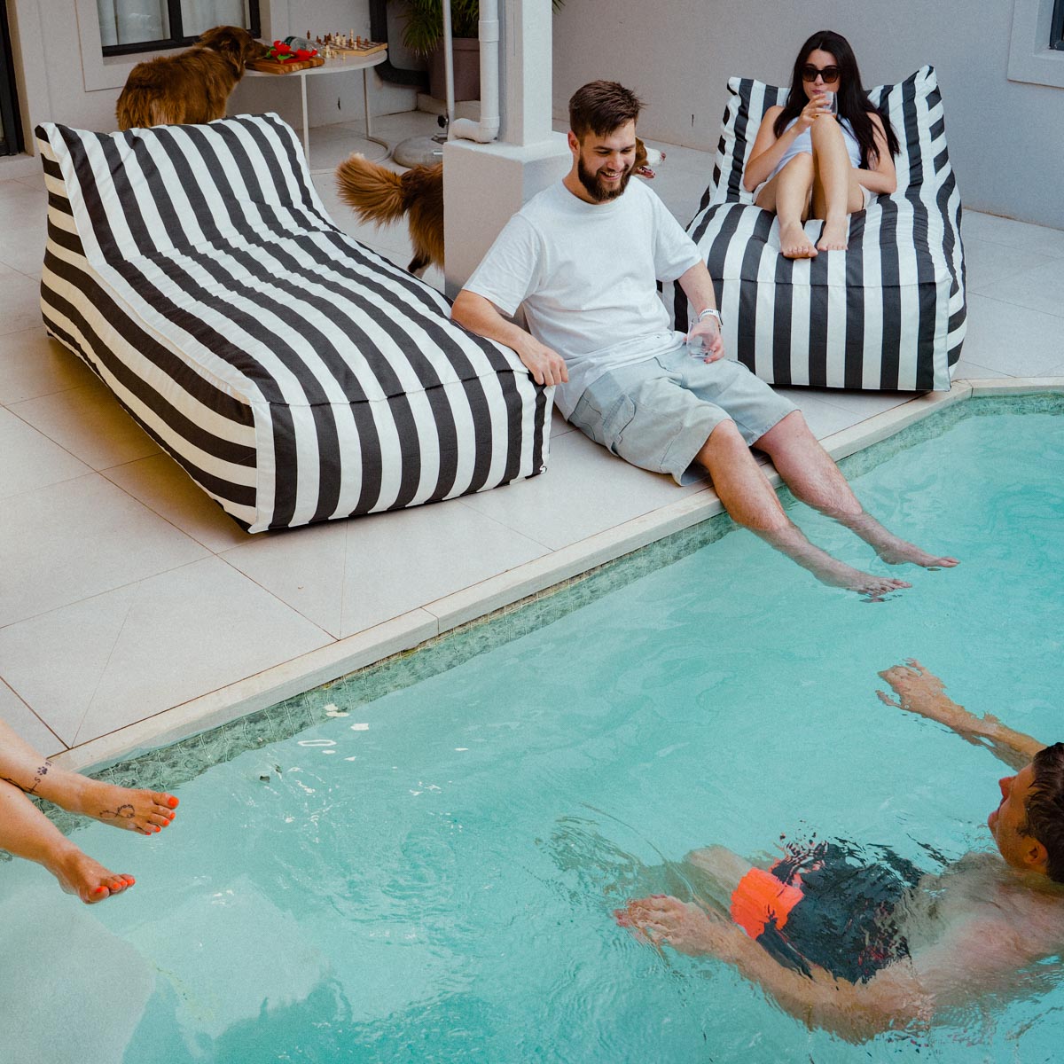 People relaxing by a pool with striped bean bags and dogs in the background