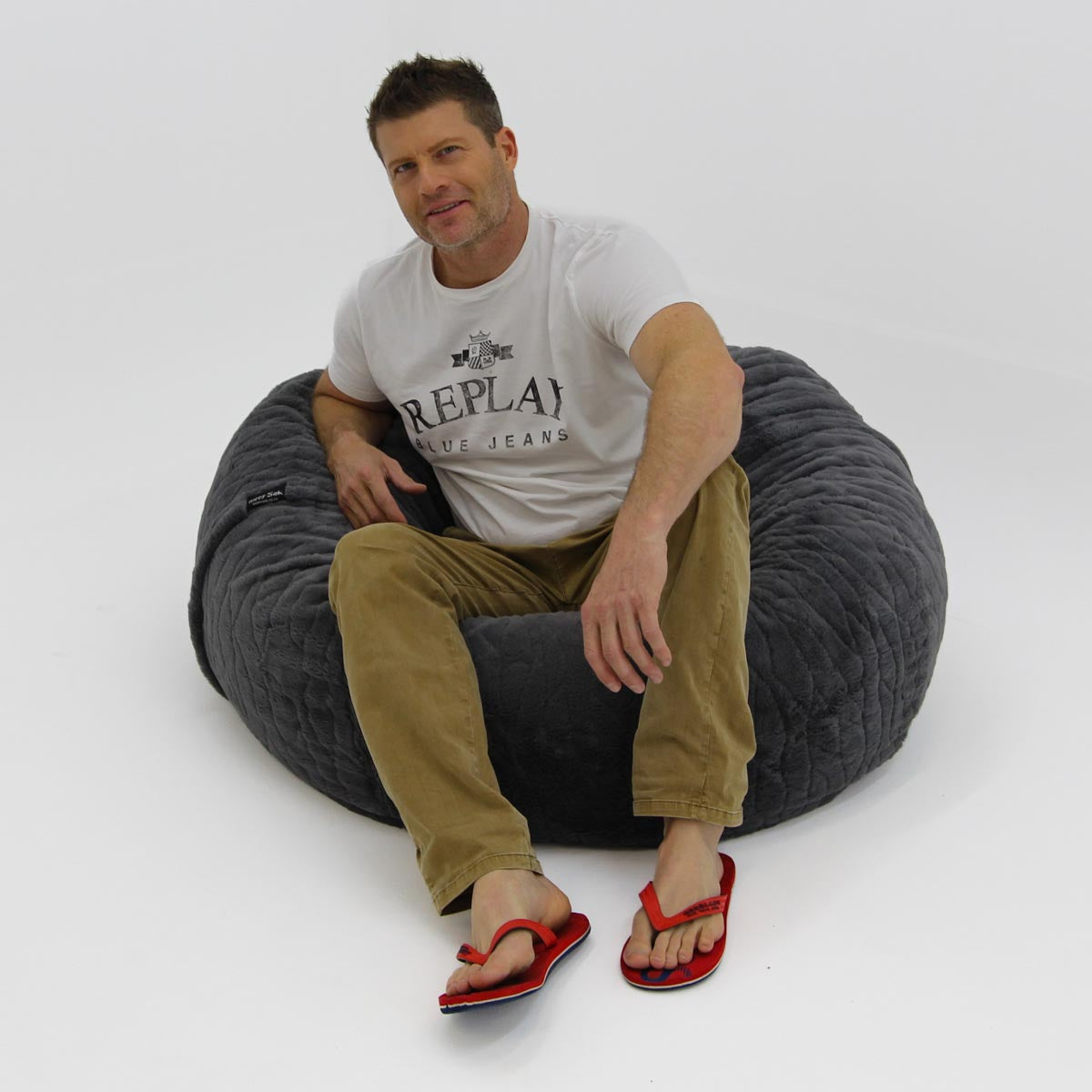 Man sitting on a large gray bean bag chair wearing a 'Replay' t-shirt.