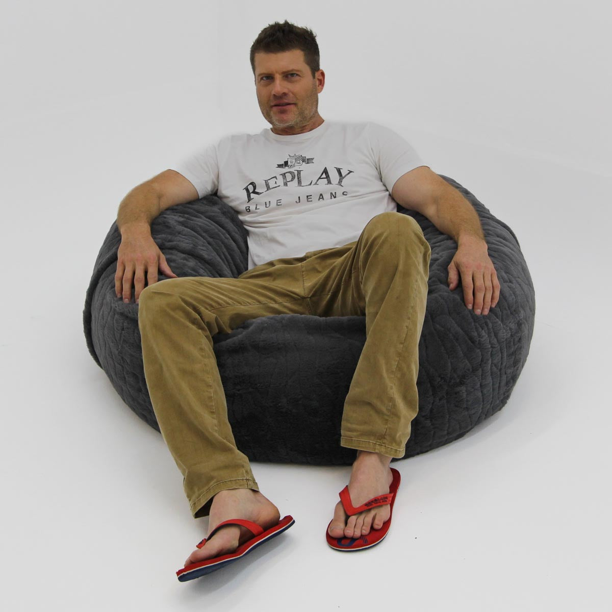 Man sitting on a gray bean bag chair wearing a white 'Replay' t-shirt and green pants.