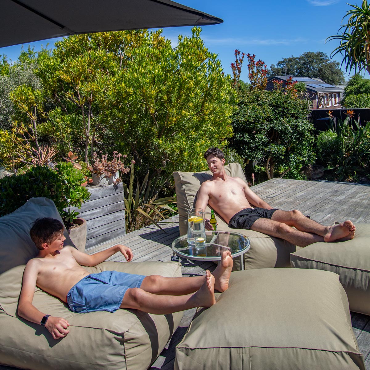 Two shirtless men lounging on beige outdoor chairs with a table between them, surrounded by greenery.