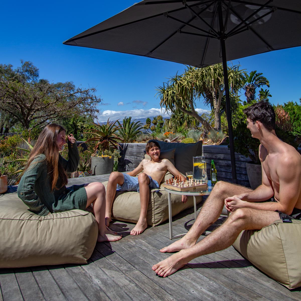 Three people sitting on outdoor furniture under a large umbrella with a scenic background.