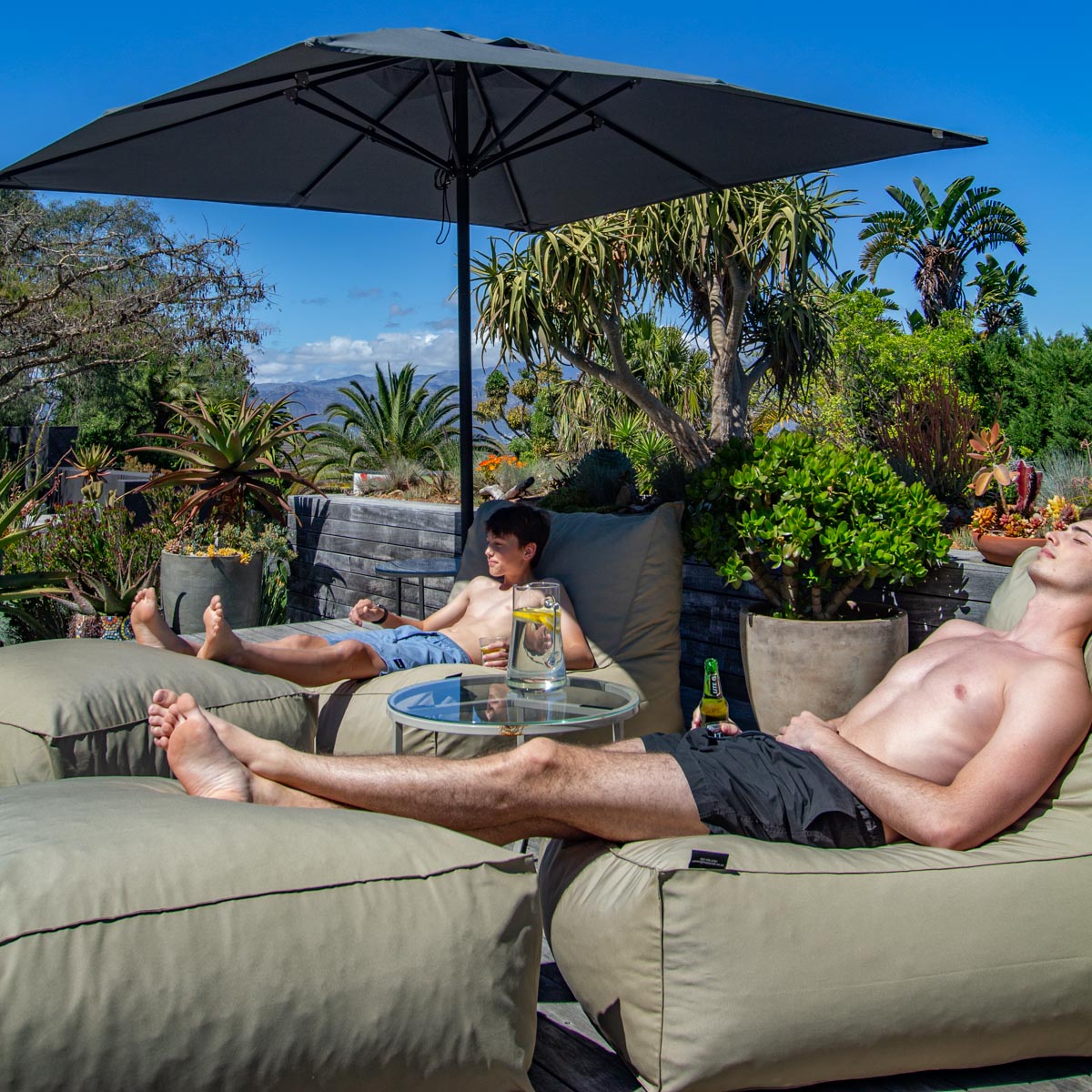 Two men relaxing on lounge chairs under a large umbrella in a garden setting.