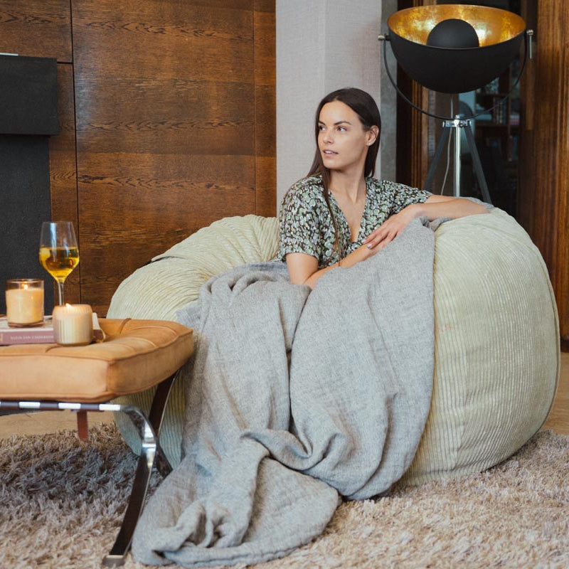 Woman sitting on a bean bag chair with a blanket, surrounded by candles and a glass of wine in a cozy room.