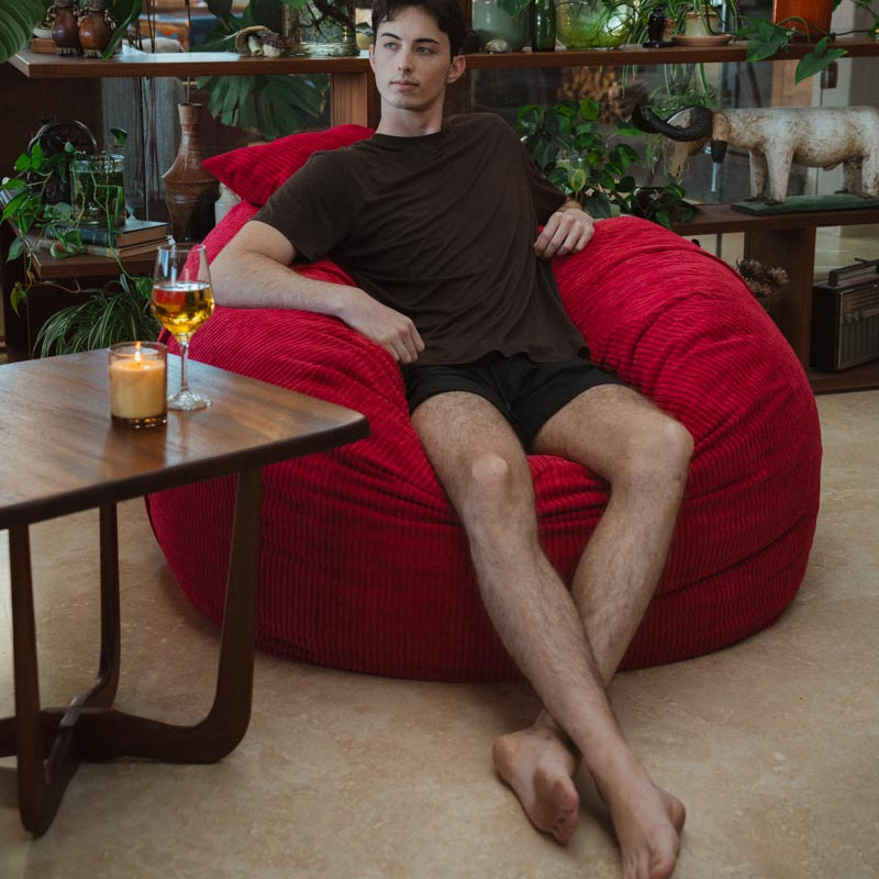 Man sitting on a red bean bag chair in a room with plants and a table.