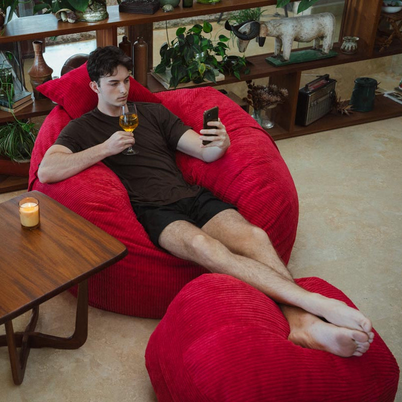 Man relaxing on a red bean bag chair holding a glass of wine and phone in a cozy living room.
