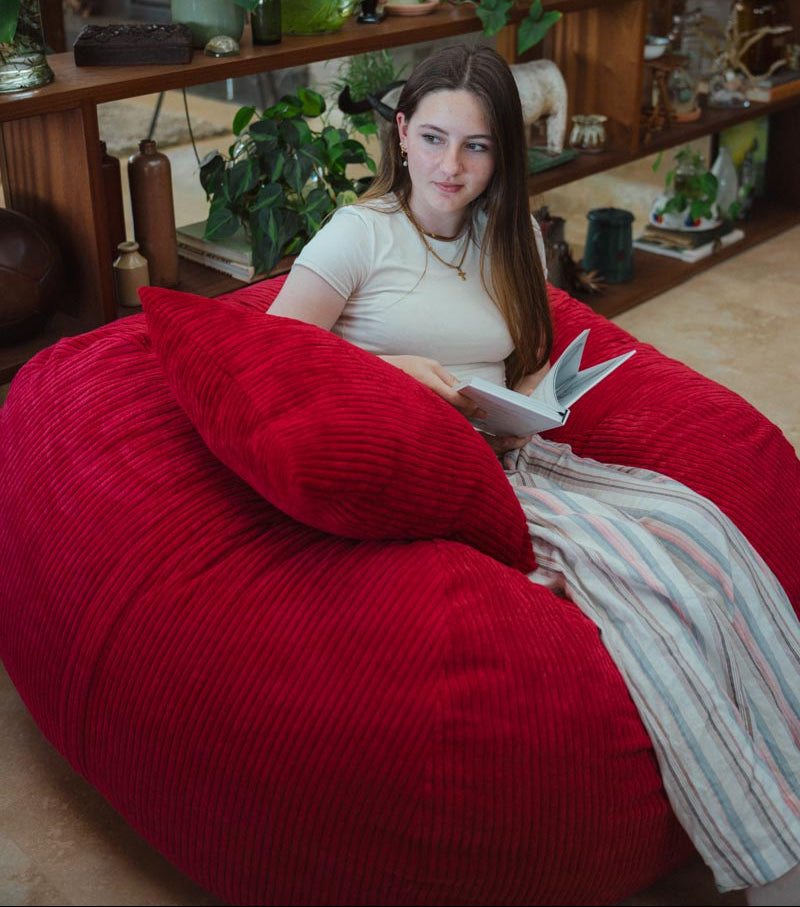 Woman reading a book on a large red bean bag chair in a cozy room.
