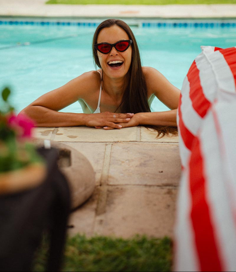 Woman wearing sunglasses and a white swimsuit, leaning on a poolside ledge with a blurred American flag in the foreground.