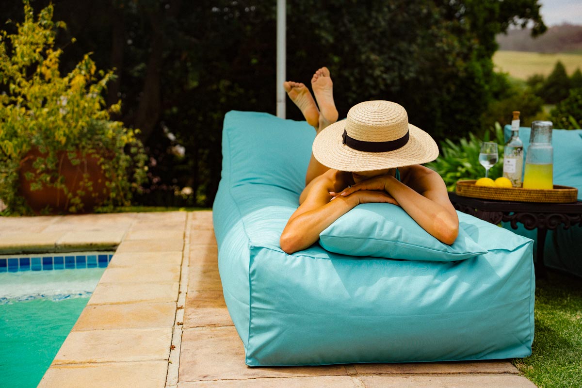Person relaxing on a blue bean bag by a poolside with a hat on