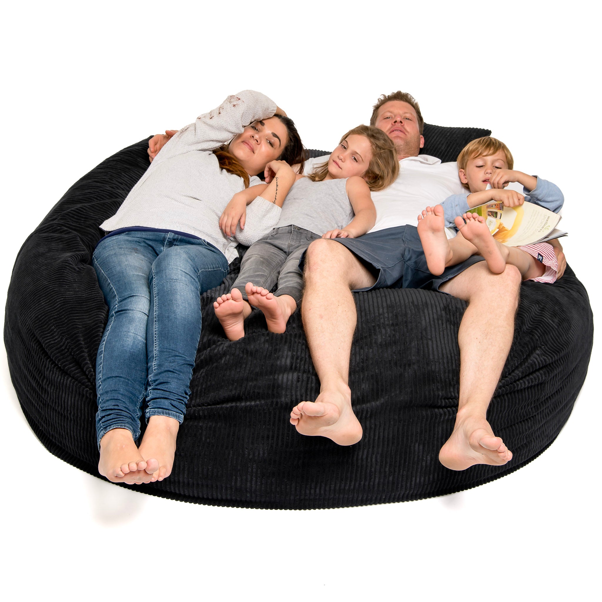 A large black corduroy bean bag, known as a 'Sak', with three adults and a child sitting and lying on it, clearly designed for comfort and relaxation.