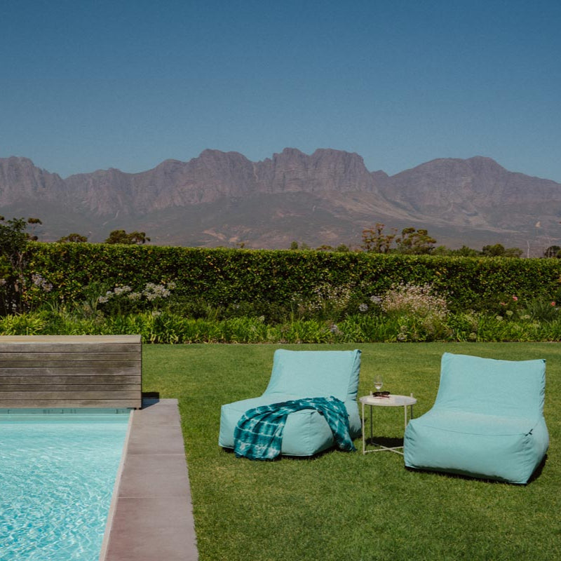 Two turquoise bean bags on grass near a pool with mountains in the background
