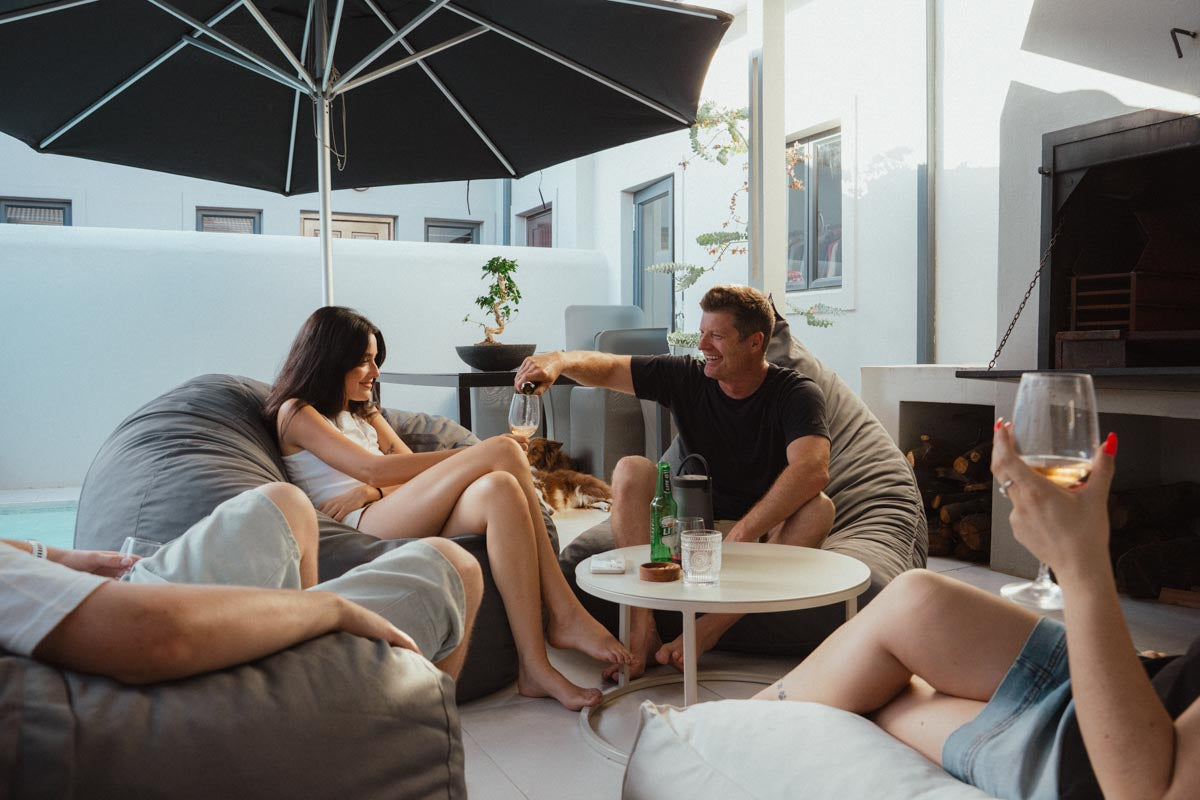 People relaxing on outdoor furniture under an umbrella with a pool and house in the background