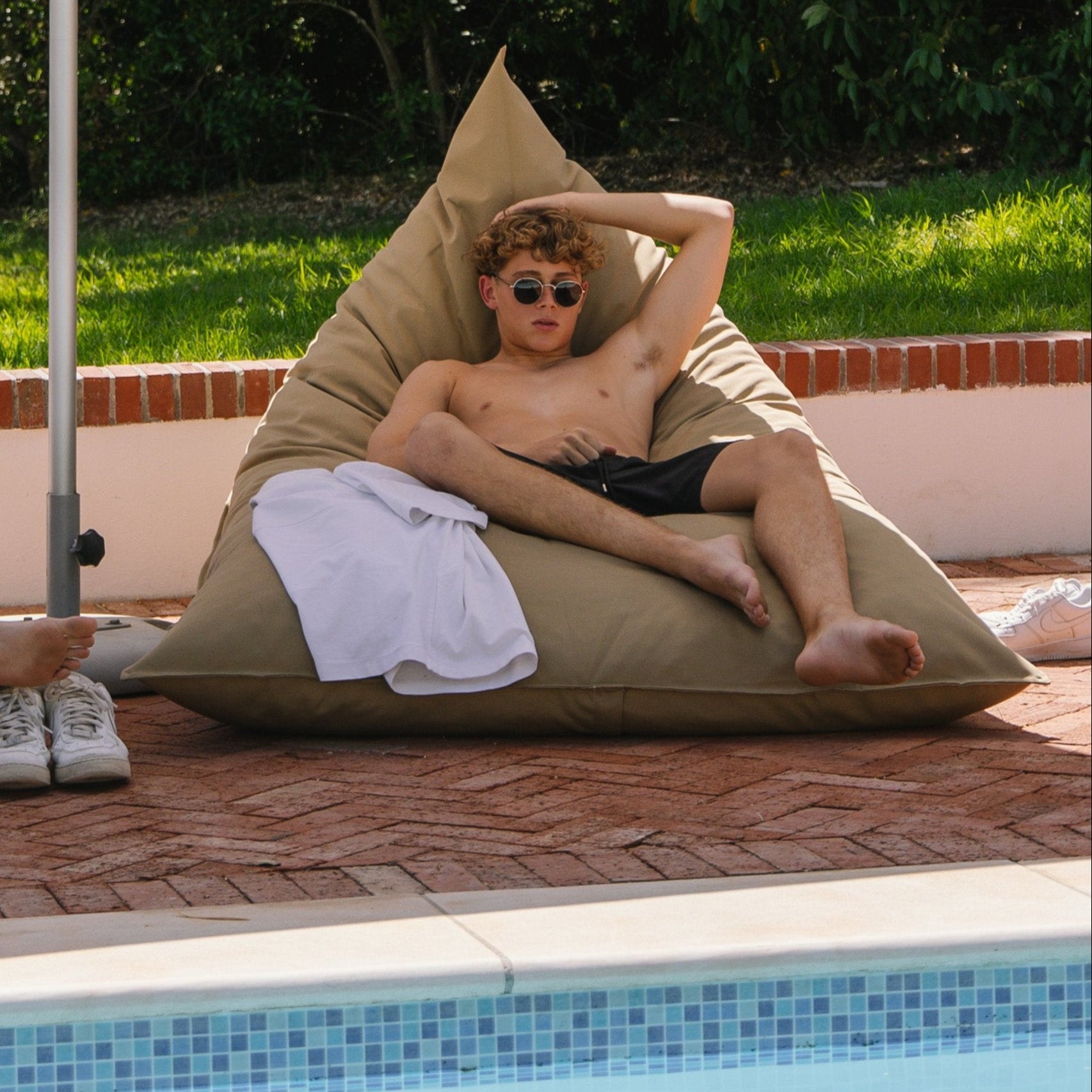 Two individuals relaxing on large beige outdoor cushions by a poolside, with a sun umbrella overhead.