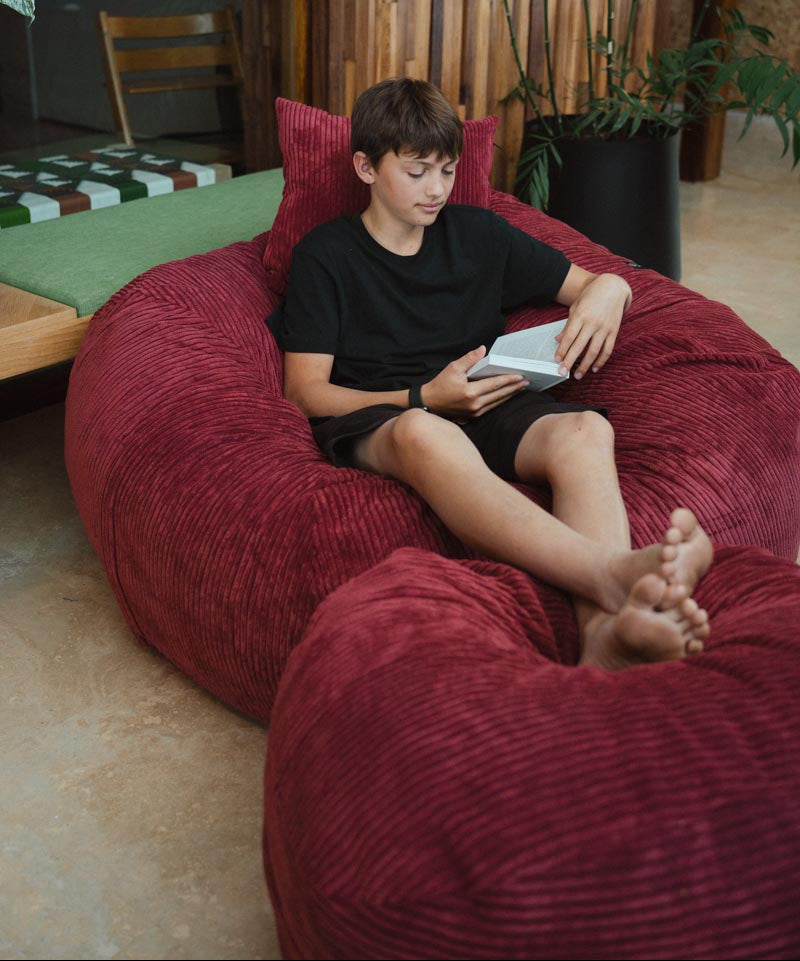 Child sitting on a large red bean bag reading a book in a casual indoor setting.