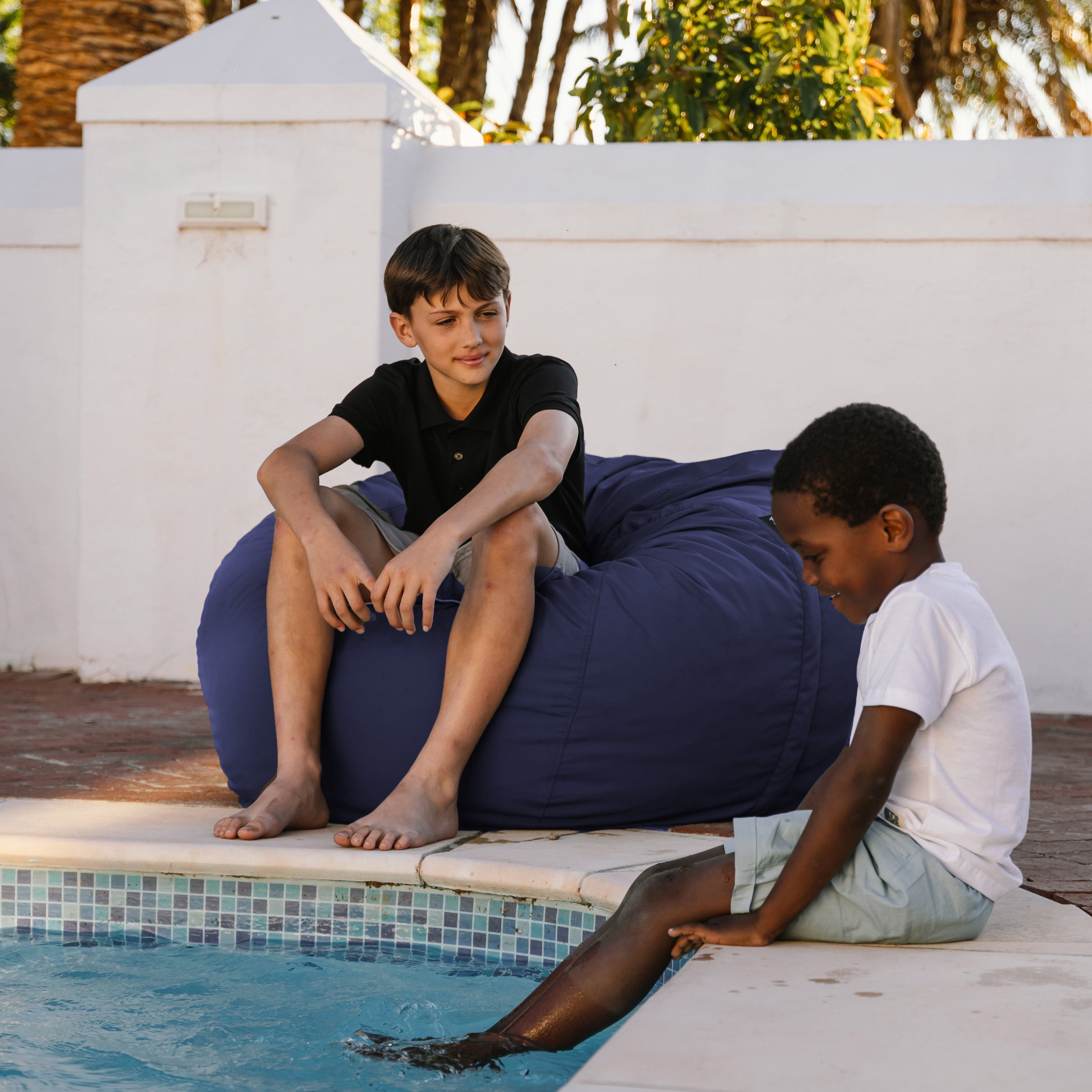 Two children sitting on a blue bean bag by a pool with palm trees in the background.