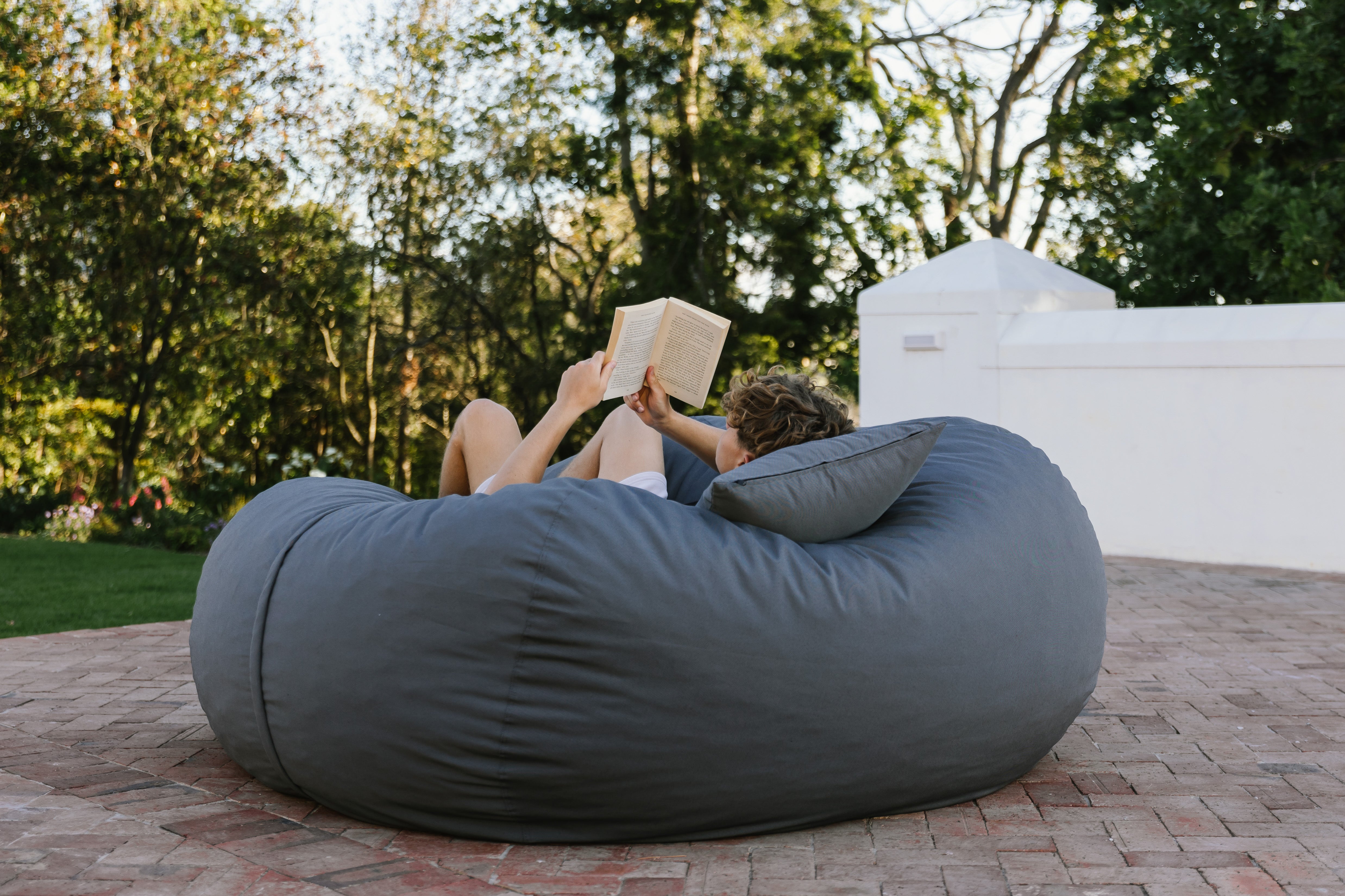 Person reading a book on a large gray bean bag chair outdoors.