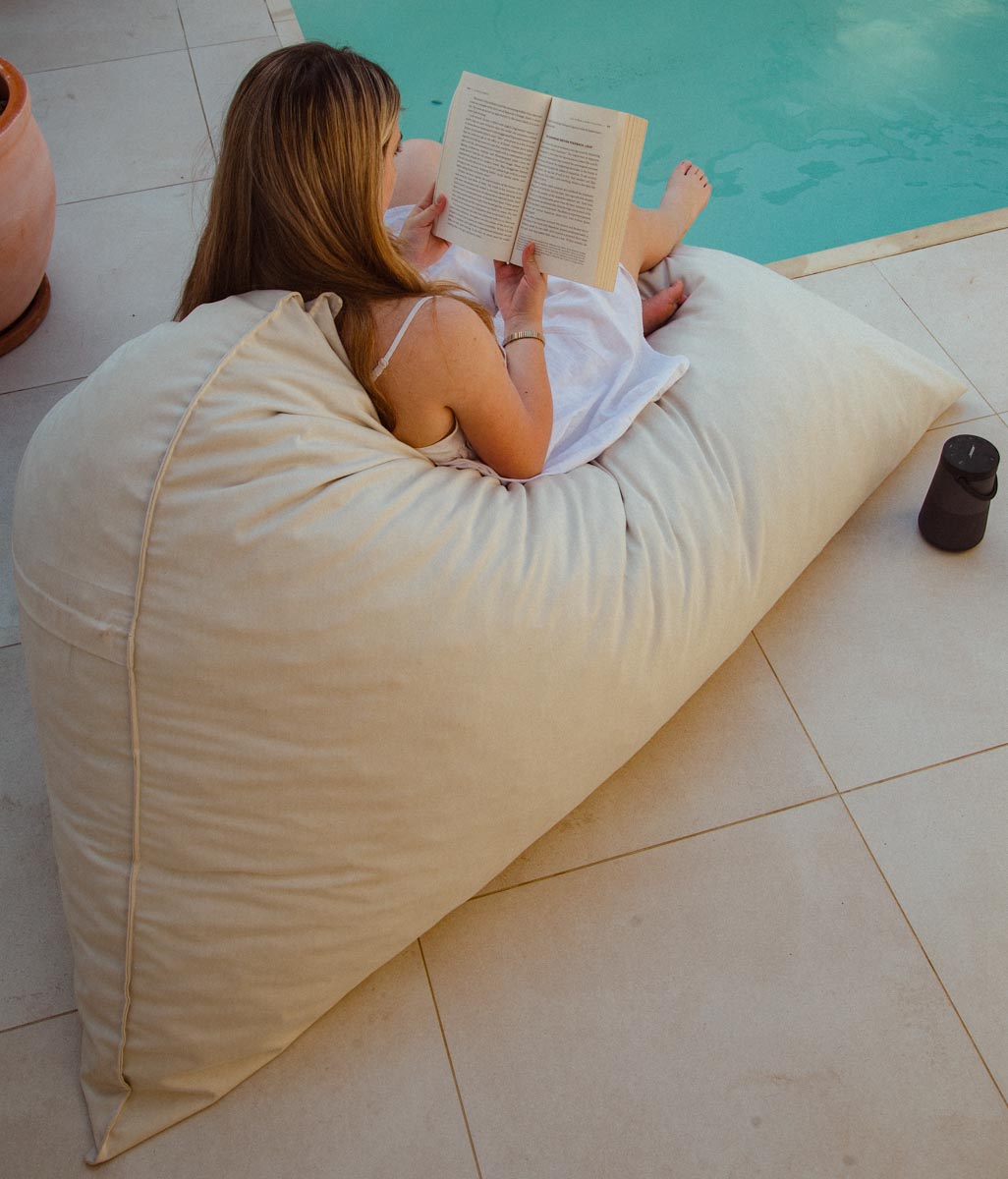 Woman reading a book on a large beige pillow by a poolside.