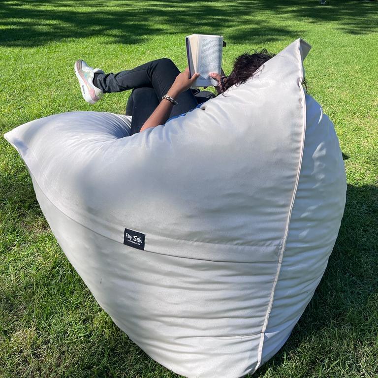 A person sitting on a gray Flip Sak outdoor bean bag chair on grass, reading a book.