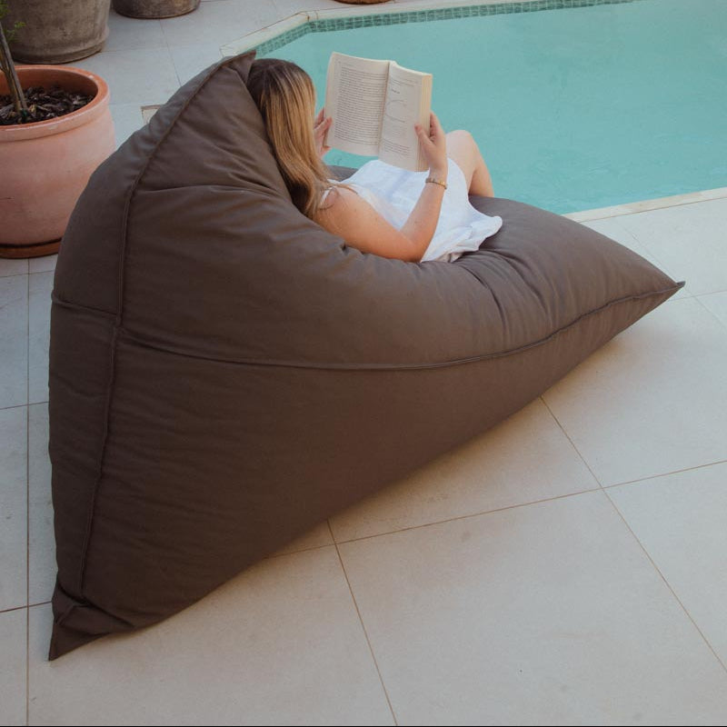 Person reading a book on a large brown bean bag by a pool with potted plants around.