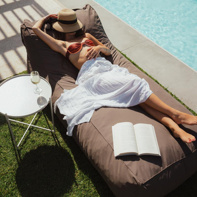 Person relaxing on a lounge chair by a pool with a book and sunglasses.