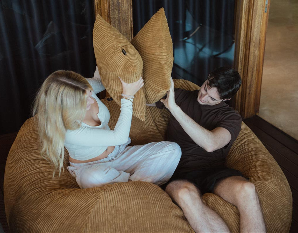 Two people playing with pillows on a brown couch.