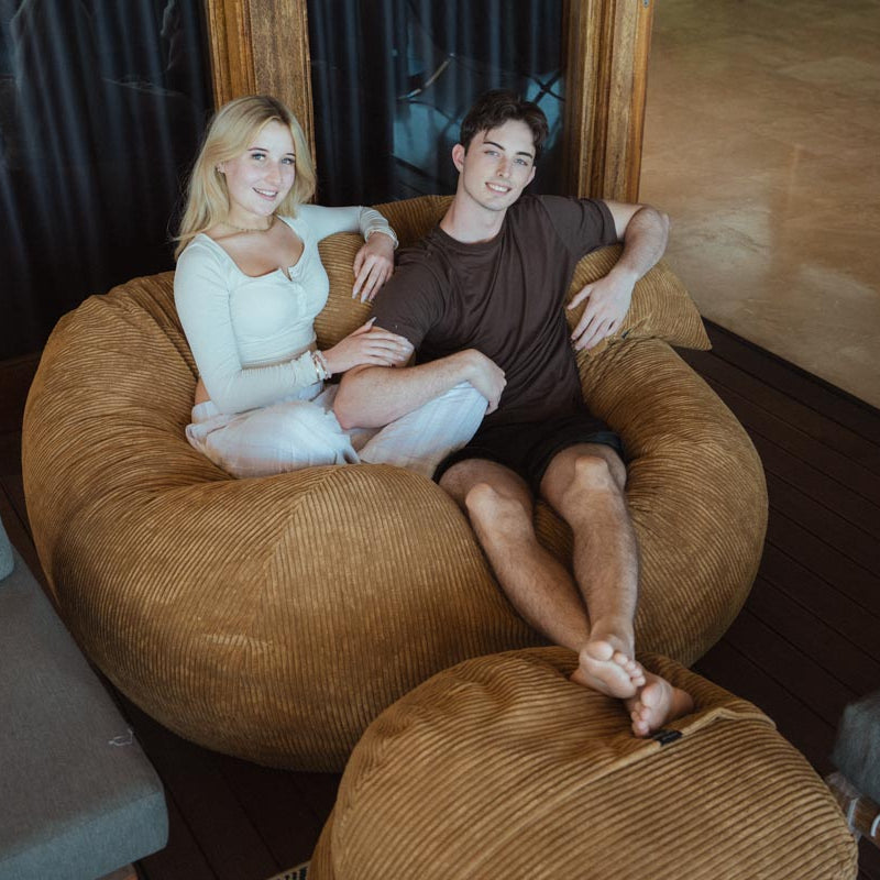 Two people sitting on a large brown bean bag chair in a cozy room.