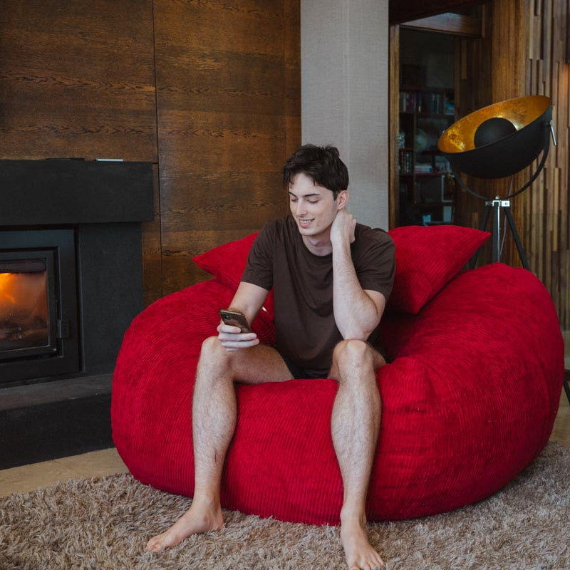 Man sitting on a red bean bag chair in a cozy living room with a fireplace.