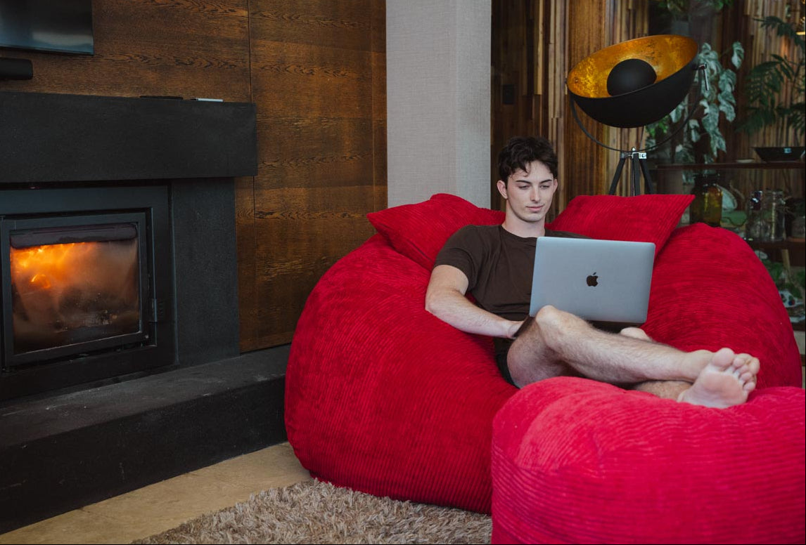Person using a laptop on a red bean bag chair in a cozy living room with a fireplace.