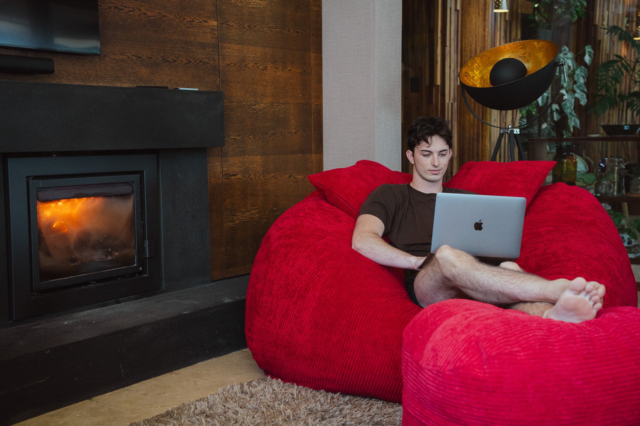 Person using a laptop on a red bean bag chair in a cozy living room with a fireplace.