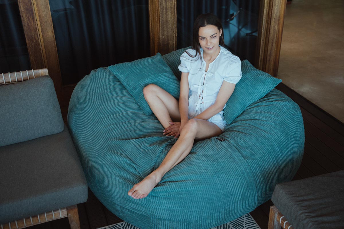 Woman sitting on a large teal bean bag chair in a room with wooden walls.