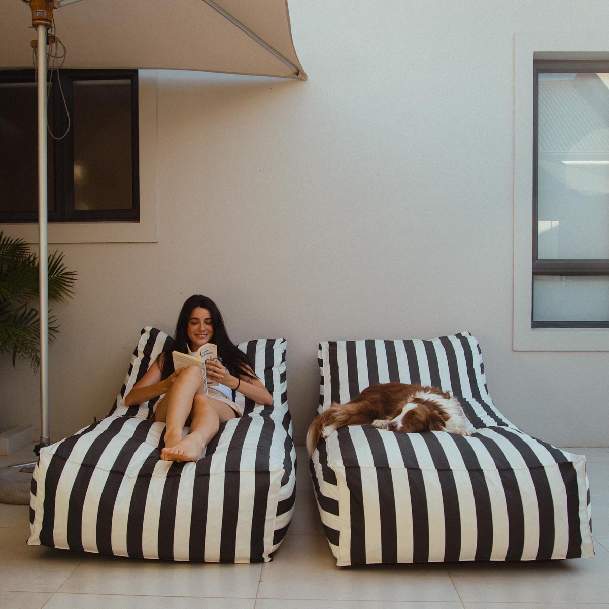 Woman sitting on a black and white striped bean bag chair reading a book, with a dog lying next to her.