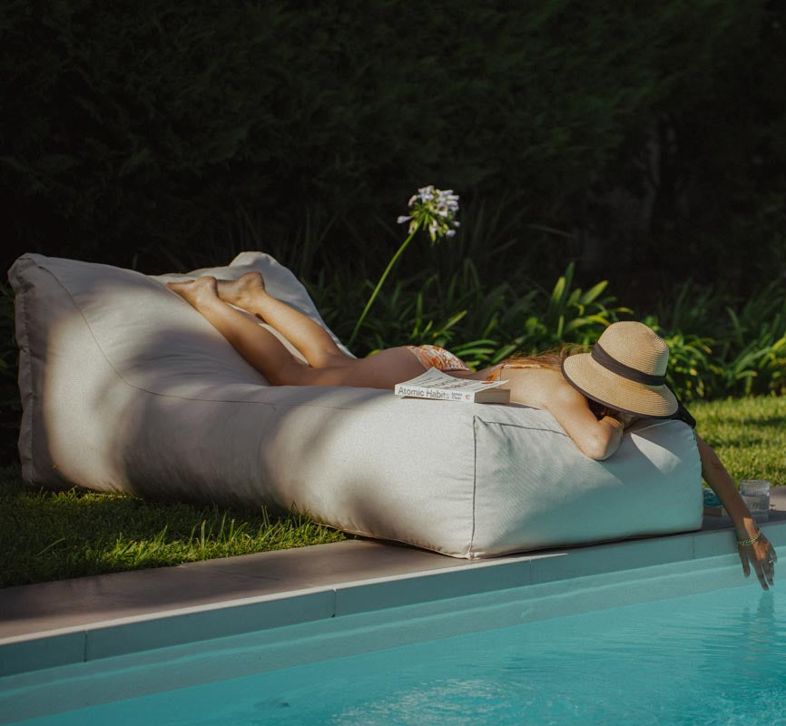 Person relaxing on a lounge chair by a pool with a book and hat.