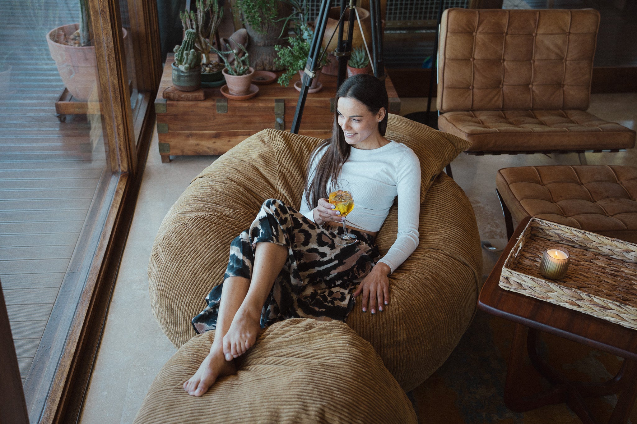 A woman relaxes in a large brown corduroy lounge chair inside a cozy room, holding a glass of yellow drink. She is wearing a white long-sleeve top and leopard-print pants.